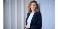 Frau mit lockigem Haar im Blazer lächelt und lehnt an einer Wand vor hellem Vorhang.