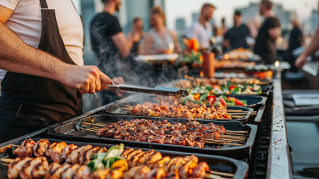 Ein Mann steht auf einem Event an einem langen Grill mit mehreren Grillschalen mit Gemüse und Fleischspießen.
