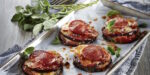 Baked eggplant rounds topped with cheese and pepperoni on a parchment-lined tray with fresh herbs alongside.