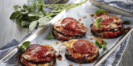 Baked eggplant rounds topped with cheese and pepperoni on a parchment-lined tray with fresh herbs alongside.