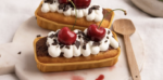 Two rectangular pastries topped with whipped cream, chocolate shavings, and cherries on a white plate with a wooden spoon nearby—dessert ready to serve.