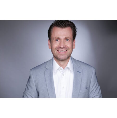 Professional head-and-shoulders portrait of a smiling man in a light gray blazer and white shirt against a neutral backdrop.