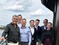 Symporo stärkt Gemeinschaftsverpflegung im Raum Lüneburg durch Übernahme von die feinschmeckerei Seven professionals pose on a river deck beside a large boat, smiling with city and cloudy sky behind.