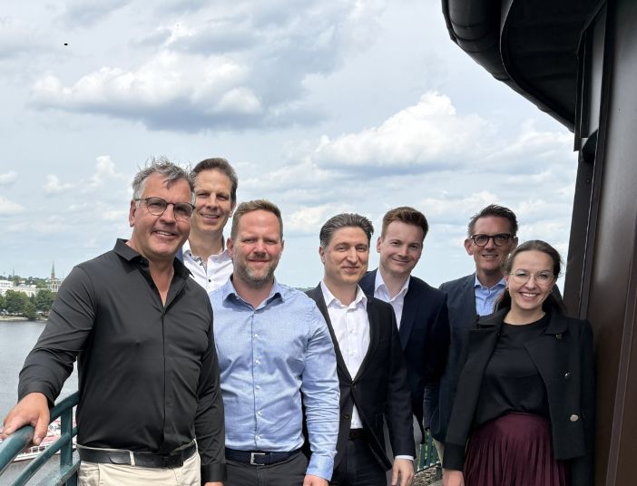Symporo stärkt Gemeinschaftsverpflegung im Raum Lüneburg durch Übernahme von die feinschmeckerei Seven professionals pose on a river deck beside a large boat, smiling with city and cloudy sky behind.