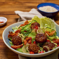 Bowl of salad with glazed meatballs, cucumber, onions, lettuce leaves, and red chili slices on a wooden table.