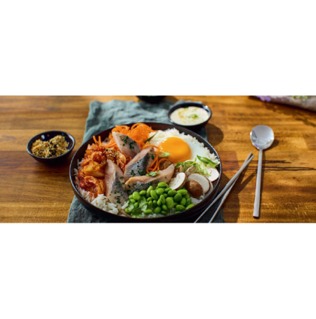 Matjes around the world – Südkorea Bowl of bibimbap with rice, vegetables, sliced beef, an egg yolk, and green peas, with chopsticks nearby on a wooden table.