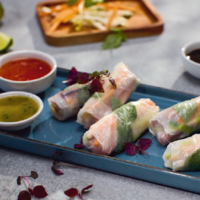 Three fresh vegetable spring rolls wrapped in translucent rice paper on a blue platter, with dipping sauces and herb garnish nearby.