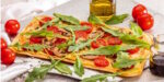 Flatbread pizza topped with arugula, mushrooms, and tomato slices, with a bottle of olive oil nearby on a rustic table.