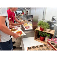 World Rowing Championships: 14.500 Sportlermahlzeiten Person in a red shirt serves themselves at a buffet station with sliced meats and cheeses under a tent.
