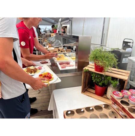 World Rowing Championships: 14.500 Sportlermahlzeiten Person in a red shirt serves themselves at a buffet station with sliced meats and cheeses under a tent.