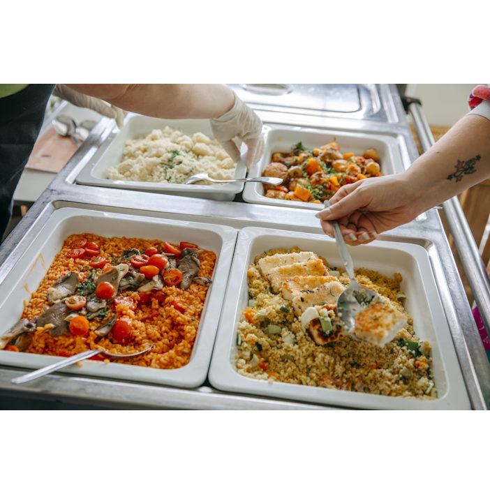 Bayerischer Schulverpflegungskongress 2026 in Nürnberg Buffet line with hands serving food from chafing dishes, including tomato-based rice, couscous, and mixed vegetables.