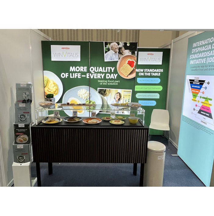 Exhibition booth with a dark wooden counter displaying plated food samples in front of a green banner reading 'More quality of life – every day'.